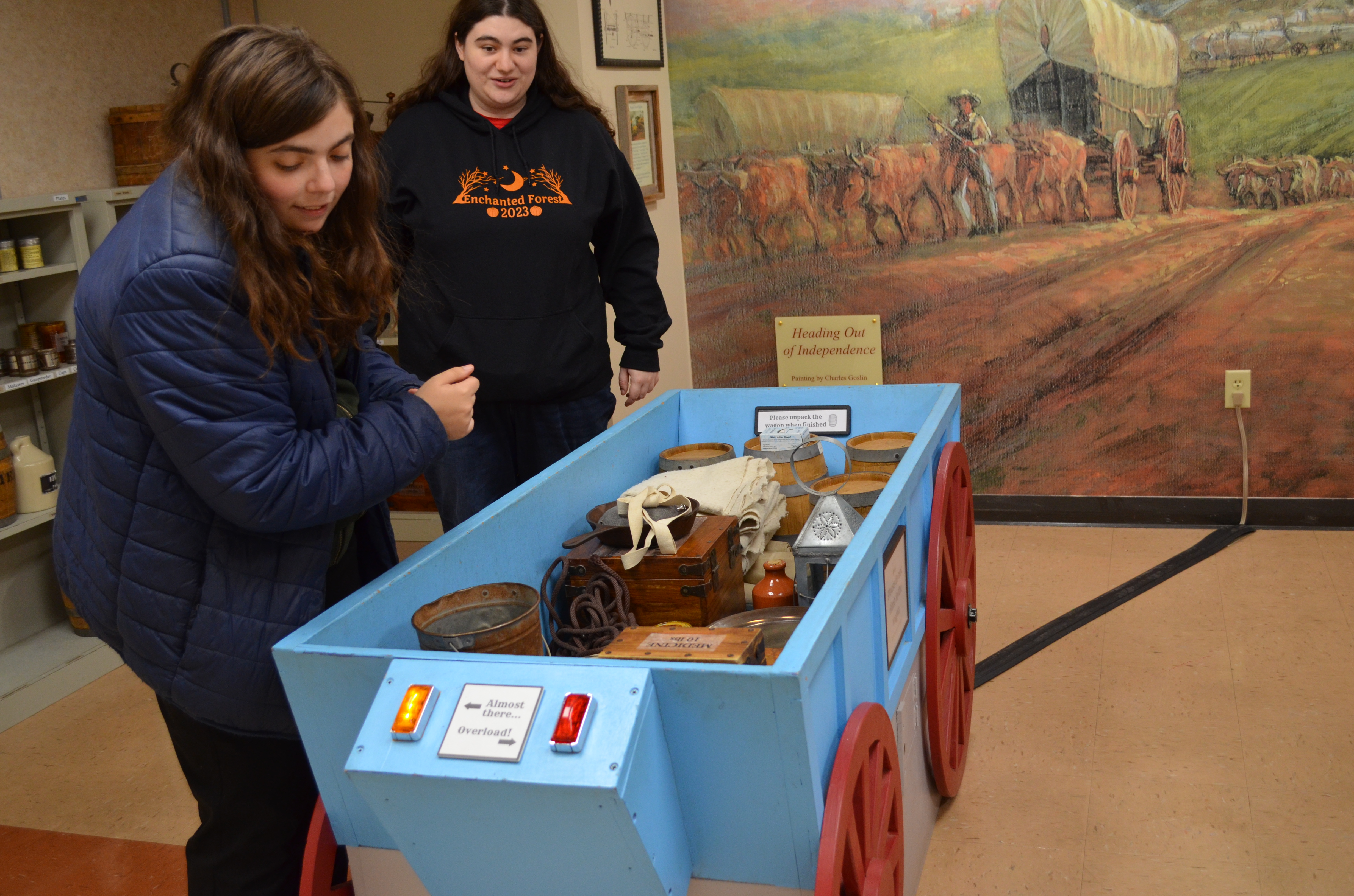 Two young women load items into a blue box representing a wagon at the National Frontier Trails Museum.