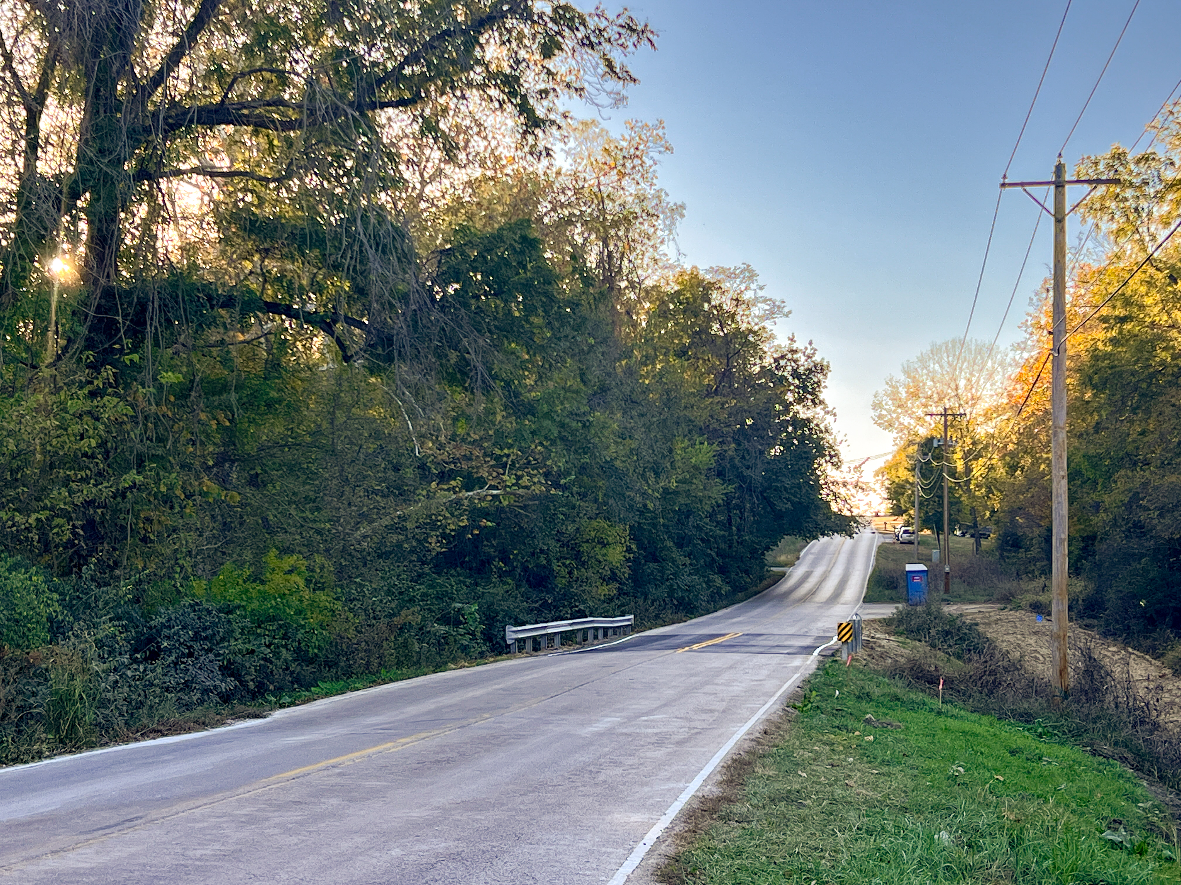 Straight paved road surrounded by trees and power lines on the right, with newly installed guardrails and a fresh pave job over a culvert that was damaged by flooding rains.