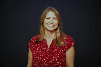 A woman sits for a portrait with a red shirt and a dark background.