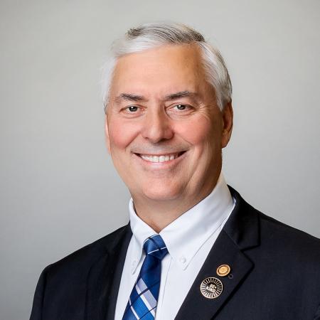 Mayor Rory Rowland smiles in portrait photo. He is wearing a dark suit, white shirt, blue, grey and white plaid tie while seated in front of a light grey background.