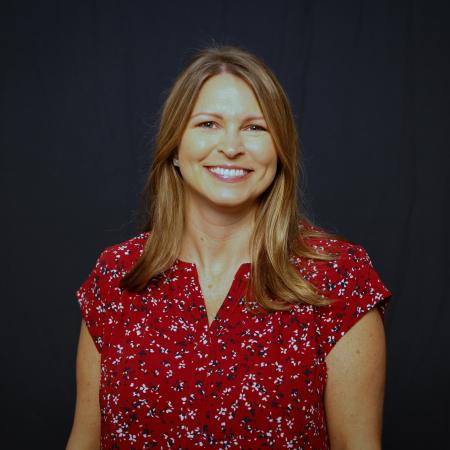 A woman sits for a portrait with a red shirt and a dark background.