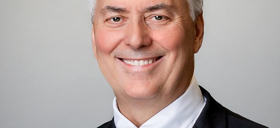 Mayor Rory Rowland smiles in portrait photo. He is wearing a dark suit, white shirt, blue, grey and white plaid tie while seated in front of a light grey background.