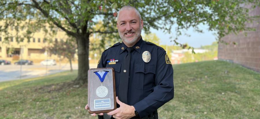 Officer Travis Gillihan stands in uniform front of a tree at sundown holding the Susan Paxton Block Award