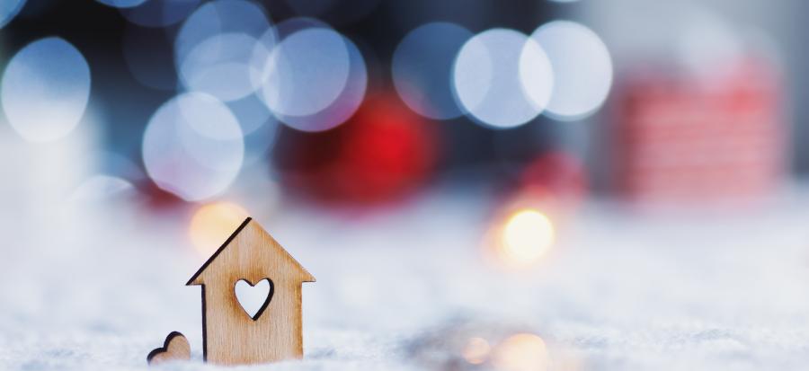 A small wooden home cutout sits in the snow with a heart cut out of the middle of it. Holiday lights and gifts are blurred in the background.