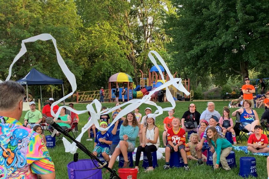 Families enjoying the entertainer in front of them during the event, Popsicles in the Park, with a playground in the background at Mill Creek Park.