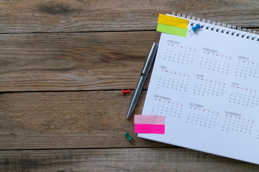 A calendar sits on a wood desk next to a pen with post-it tabs placed on one side. 