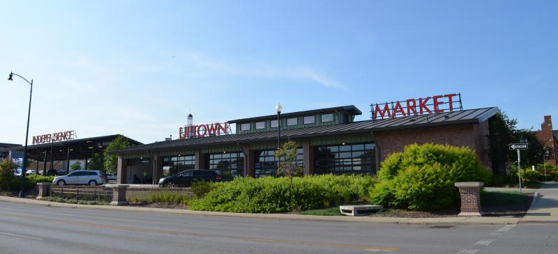 An image of the north side of the Independence Uptown Market building during the daytime with a few parked vehicles and some green landscape.