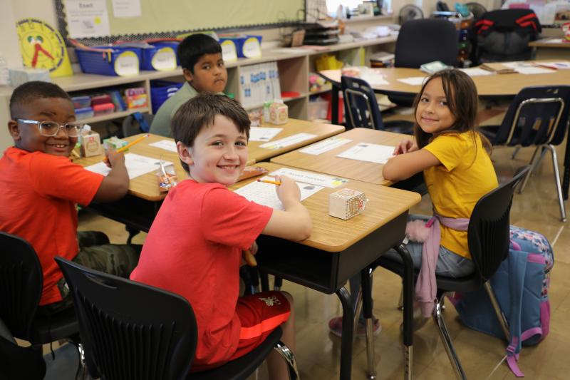Four students turn to look at the camera from their desks in an elementary school classroom.