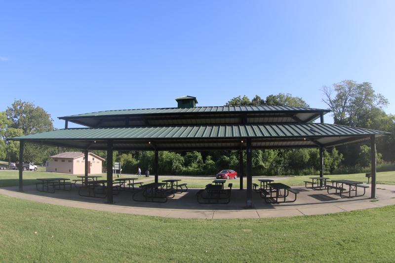 An image of the pavilion structure with 12 picnic tables and 3 grills at Hill Park with the restroom structure in the background.