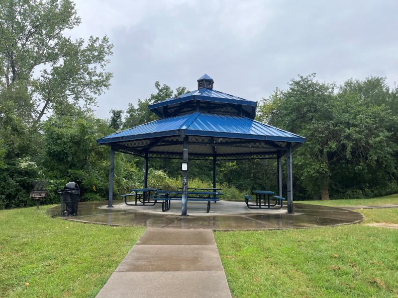 An image of a sidewalk leading up to a small round shelter with picnic tables at Mill Creek Park. Trees sit in the background.