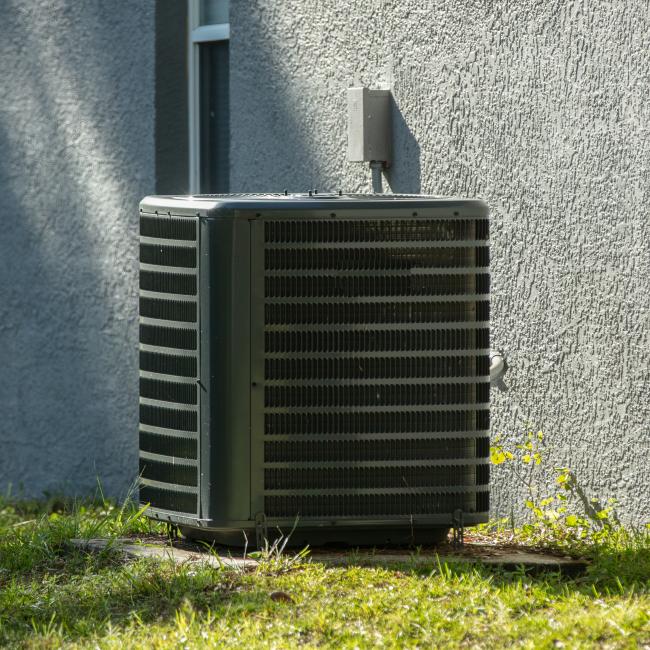 A dark grey air conditioner on a cement pad in front of a grey stucco wall at a home.