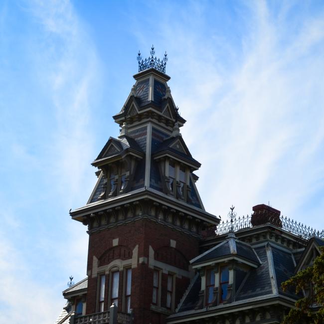 The peaked roof of the historic Victorian Vaile Mansion stands in the middle of the image with wispy white clouds in a blue sky behind it. 